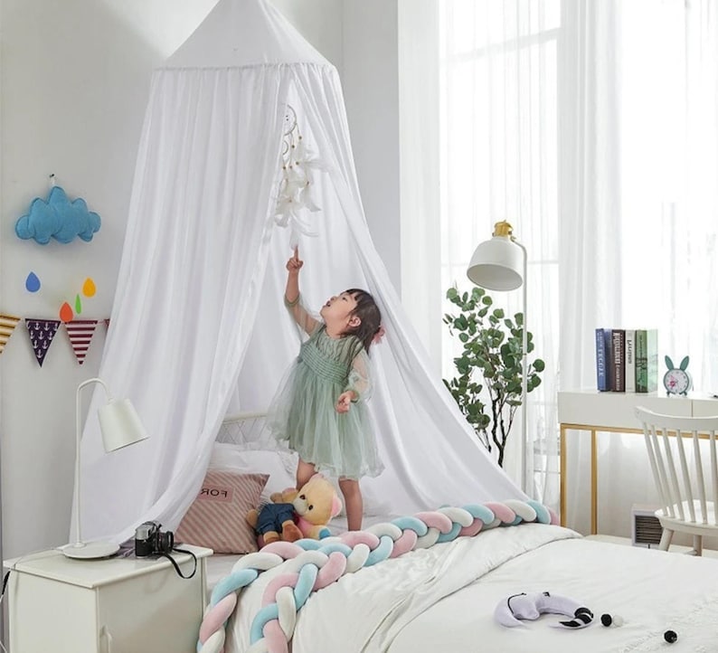 May include: A white canopy bed with a sheer, draped design. A young child in a green dress stands on the bed, reaching towards a hanging mobile. The bed is decorated with a braided bumper in pastel colors.