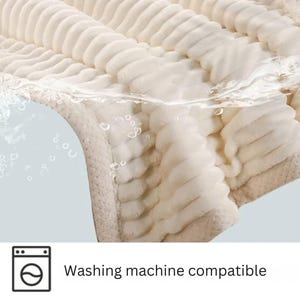 May include: A close-up of a cream-colored, plush blanket with a ribbed texture. The blanket is partially submerged in water, with bubbles visible. The text "Washing machine compatible" is displayed below a washing machine icon.