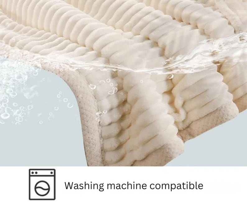 May include: A close-up of a cream-colored, plush blanket with a ribbed texture. The blanket is partially submerged in water, with bubbles visible. The text "Washing machine compatible" is displayed below a washing machine icon.