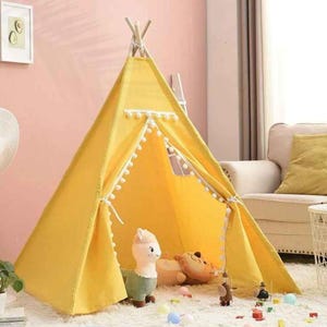 May include: A bright yellow teepee tent with white pom-pom trim and a small window. Inside the tent are stuffed animals and toys. The tent is set up on a white rug in a room with a couch and a plant.