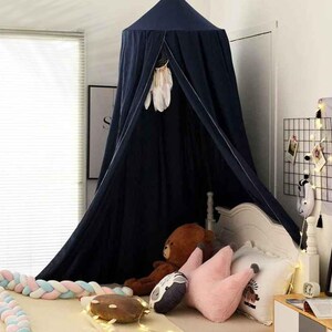 May include: A navy blue canopy bed with a dreamcatcher hanging inside. The bed is decorated with pillows, a teddy bear, and a braided bumper. A string of lights and framed art are also visible.