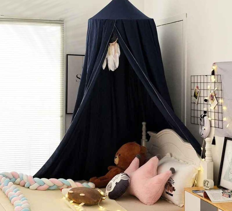 May include: A navy blue canopy bed with a dreamcatcher hanging inside. The bed is decorated with pillows, a teddy bear, and a braided bumper. A string of lights and framed art are also visible.