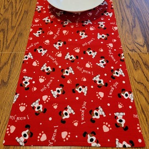 May include: Red table runner with a repeating pattern of white dogs with black noses and brown ears. The dogs are surrounded by white paw prints and pink hearts. The text "I woof you" and "I woof you" is repeated throughout the pattern.
