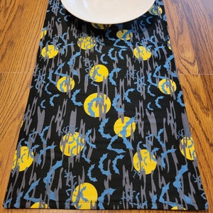 May include: Black table runner with a repeating pattern of blue bats and yellow moons. The runner has a stitched edge.