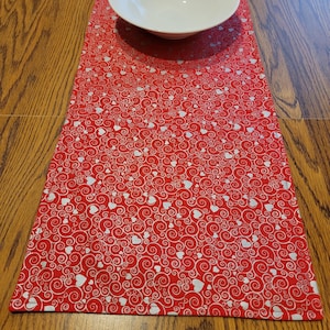 May include: Red table runner with a white swirl and heart pattern. The runner is on a wooden table with a white bowl on top.