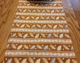 May include: A brown and white table runner with a repeating pattern of oak leaves and acorns. The runner is placed on a wooden table and has a white bowl at the top.