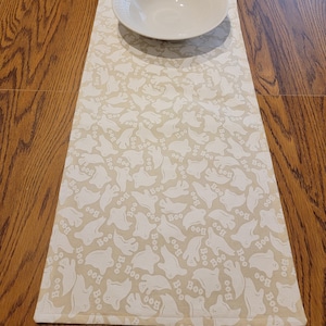 May include: A beige table runner with a white ghost pattern and the word "Boo" repeated throughout.