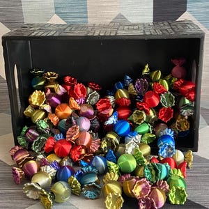 May include: A black wooden crate filled with colourful, wrapped sweets. The sweets are in various colours, including red, blue, green, and gold. The sweets are scattered on a geometric patterned surface.