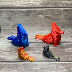 May include: Four colorful cardinal bird figurines in blue, red, orange, and black. The birds are perched on a wooden surface. The figurines have detailed wings and crests, and are a decorative item.