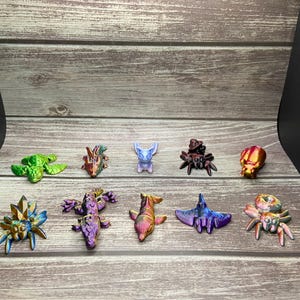 May include: A collection of ten miniature fantasy creatures in various colours. The creatures include a turtle, dragon, deer, spider, and a skull. The figurines are arranged on a wooden surface.