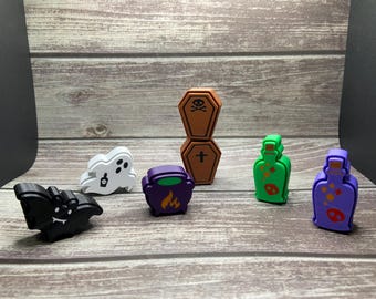 May include: A collection of six small Halloween-themed figurines including a black bat, a white ghost, a purple cauldron, two stacked brown coffins with skull and cross symbols, a green potion bottle, and a purple potion bottle with skull icons.