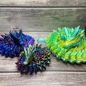 May include: Three crystal dragon sculptures in shades of blue, green, and iridescent colors. The dragons are posed on a wooden surface. The sculptures have a spiky, geometric design, resembling crystals.