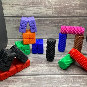 May include: A collection of colorful fidget toys in various shapes and sizes. The image features cubes, cylinders, and articulated blocks in shades of purple, orange, green, blue, black, pink, brown, and red. The toys are arranged on a wooden surface.