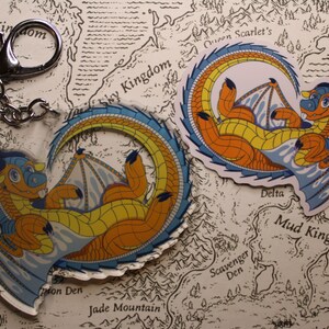 Puede incluir: Dos llaveros redondos de acr&iacute;lico transparente con un drag&oacute;n de dibujos animados con escamas azules y naranjas. El drag&oacute;n est&aacute; enroscado en un c&iacute;rculo con las alas extendidas. Los llaveros est&aacute;n en un mapa con las palabras "Ice Kingdom", "Sky Kingdom", "Mud Kingdom", "Scavenger Den", "Jade Mountain" y "Delta".