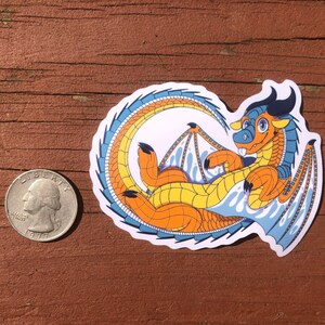 Puede incluir: Una pegatina de un drag&oacute;n de dibujos animados con escamas naranja, amarillo y azul. El drag&oacute;n est&aacute; enroscado con las alas extendidas. La pegatina est&aacute; sobre una superficie de madera.