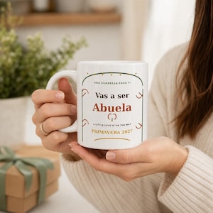May include: A white ceramic mug featuring the text "UNA SORPRESA PARA TI", "Vas a ser Abuela", "A LITTLE LOVE IS ON THE WAY", and "PRIMAVERA 2027" in black and brown lettering, framed by a thin green arch design with small dots.