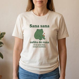 May include: A cream-colored short-sleeve t-shirt featuring a green frog illustration with a small orange bandage on its leg and a red heart. The text reads: Sana sana colita de rana, si no sana hoy, sanará mañana, printed in dark green lettering.