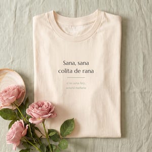 May include: A light beige crew neck t-shirt featuring the black printed text Sana, sana colita de rana with a horizontal line underneath and the smaller grey text si no sana hoy, sanará mañana centered on the chest of the garment.