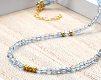 May include: A beaded necklace featuring translucent light blue round beads interspersed with small, polished gold-toned metal spacers. The necklace includes a gold-toned metal lobster claw clasp and an adjustable chain extender at the end.