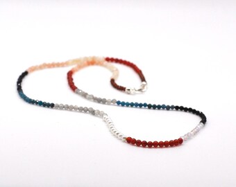 May include: A beaded necklace featuring a sequence of small, round, faceted stones in various colors including red, blue, grey, black, white, and peach, finished with a silver-toned metal clasp closure at the back of the strand.
