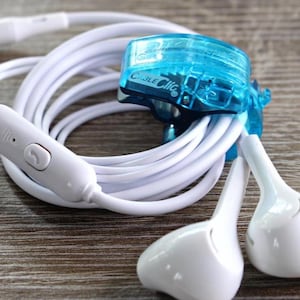 Puede incluir: Auriculares internos blancos con cable blanco, clip de cable azul y botón de control blanco. El cable está enrollado y los auriculares están sobre una superficie de madera.