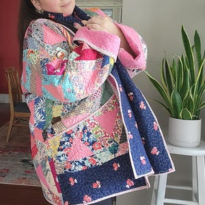 May include: A long-sleeved jacket constructed from a patchwork quilt featuring a variety of floral and geometric patterns in shades of pink, blue, and white. The garment has a dark blue interior lining with a small pink floral print and pink trim.