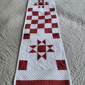 May include: A quilted table runner in white and red. The runner features a checkerboard pattern, star designs, and decorative stitching. The runner is approximately 60 inches long.