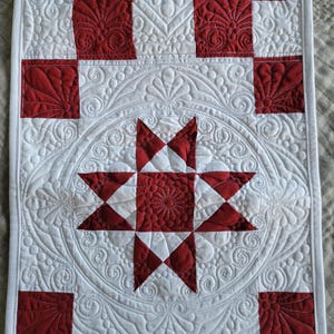 May include: A quilted table runner featuring a central red and white star design. The runner has a checkerboard pattern with red squares and white squares. The white sections have intricate quilted patterns, and the edges are finished with a white border.