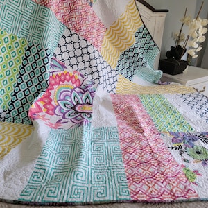 May include: A patchwork quilt featuring a variety of colorful fabric patterns including teal Greek key, pink lattice, yellow fan shapes, black and white geometric grids, and floral prints with mandala designs, all stitched together in a quilted texture.