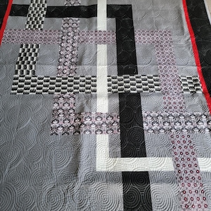 May include: A geometric quilt with a modern design. The quilt features a gray background with black, white, and patterned fabric blocks. The edges are bordered with black and a red accent. The quilting pattern includes spiral designs.
