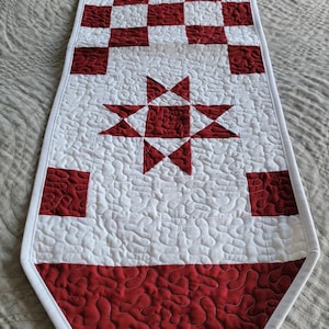 May include: A quilted table runner in red and white. The runner features a repeating pattern of red and white squares, a central star design, and a red border. The white sections have a textured, quilted pattern.