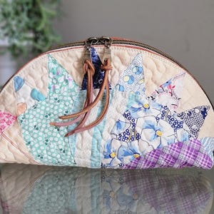May include: A quilted, half-moon shaped pouch with a zipper closure. The patchwork design features various floral and geometric patterns in shades of blue, pink, and purple. The pouch has brown leather zipper pulls.