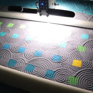 May include: A gray quilt with a swirling pattern is being quilted on a longarm quilting machine. The quilt has squares of different colors, including blue, green, yellow, and red.