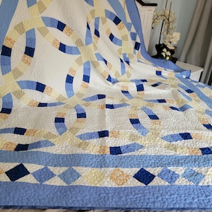 May include: A quilt featuring a double wedding ring pattern in shades of light blue, dark blue, and pale yellow on a white background. The border displays a diamond pattern in matching colors, and the entire surface has a quilted texture.