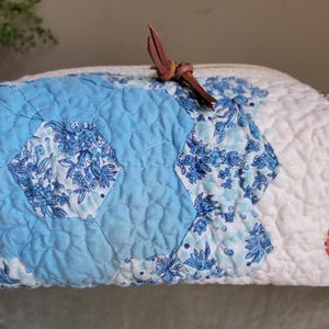 May include: A quilted zippered pouch featuring a patchwork design of hexagonal fabric pieces in shades of light blue, white, and floral patterns. The pouch is finished with a brown leather cord pull attached to the zipper slider at the top center.