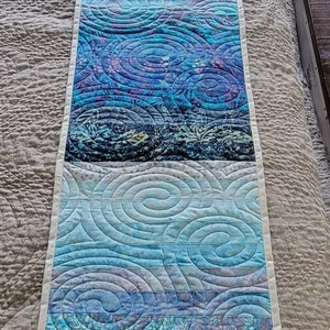 May include: A blue and purple quilted table runner with a swirling pattern. The runner is made of fabric with a batik design.