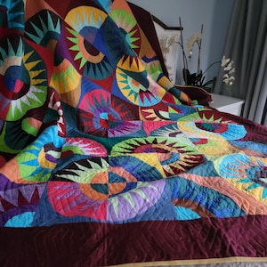 May include: A multicolored patchwork quilt featuring repeating circular sunburst patterns in shades of red, orange, yellow, green, blue, and purple, bordered by a solid dark maroon fabric with visible diagonal machine-stitched quilting lines.