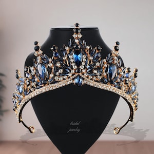 May include: A royal crown with a gold-coloured frame, embellished with sapphire blue and clear crystals. The tiara is displayed on a black mannequin head. The text "bridal jewelry" is visible on the base.