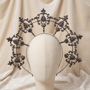 May include: A dark grey metal headpiece with an elaborate, symmetrical design. The headpiece features floral and pointed elements, attached to a thin headband. The design is displayed on a mannequin head against a neutral background.