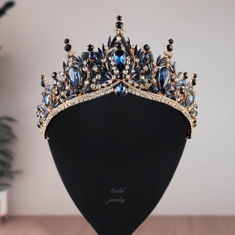 May include: A gold-toned crown with blue and clear gemstones. The crown features large, oval-shaped blue stones and smaller, round black stones. The base of the crown is lined with clear crystals. The words "bridal jewelry" are written on the display.