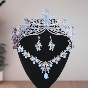 Op de afbeelding: Een zilverkleurige sieradenset bestaande uit een tiara, een ketting en een paar oorbellen. Elk sieraad is versierd met lichtblauwe, druppelvormige kristallen en heldere strass-steentjes in gedetailleerde bloem- en bladpatronen.