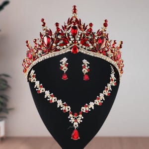 Könnte beinhalten: Ein rotes und goldfarbenes Brautschmuck-Set, bestehend aus Krone, Halskette und Ohrringen, präsentiert auf einem schwarzen Schaufensterkopf. Die Krone und der Schmuck sind mit roten Edelsteinen und klaren Kristallen verziert.
