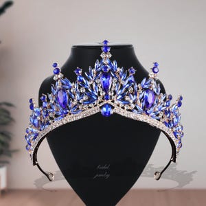 May include: A regal blue and gold tiara with sparkling blue gemstones and clear crystals. The crown is displayed on a black stand, showcasing its intricate design. The words "bridal jewelry" are written on the stand.