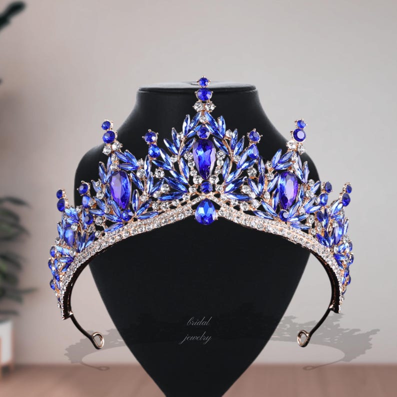May include: A regal blue and gold tiara with sparkling blue gemstones and clear crystals. The crown is displayed on a black stand, showcasing its intricate design. The words "bridal jewelry" are written on the stand.