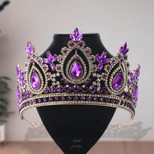 Peut inclure: Une couronne en métal doré ornée de grands cristaux violets en forme de poire et de petits strass transparents ronds. Le design présente un pic central surélevé avec un travail de filigrane complexe, accentué par des pierres violettes.