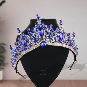 May include: A regal blue and gold tiara with large, oval-shaped blue gemstones and smaller, clear crystals. The tiara is displayed on a black stand, with the words "bridal jewelry" in script. The design features a floral pattern.