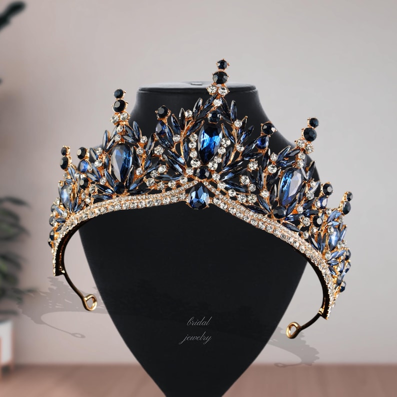 May include: A gold-toned crown with blue and clear gemstones. The crown features large, oval-shaped blue stones and smaller, clear crystals. The base of the crown is lined with clear crystals. The text "bridal jewelry" is visible on the display stand.