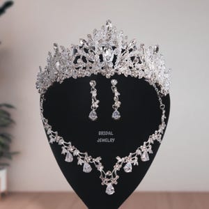 May include: A silver bridal jewelry set with a tiara, necklace, and earrings. The tiara is adorned with clear rhinestones and has a delicate floral design. The necklace and earrings are also decorated with clear rhinestones and have a similar floral design.