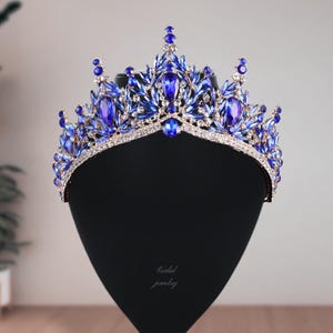 May include: A regal blue and silver crown with large, oval-shaped blue gemstones and smaller, round blue accents. The crown is set on a black display stand, with the words "bridal jewelry" in cursive script.