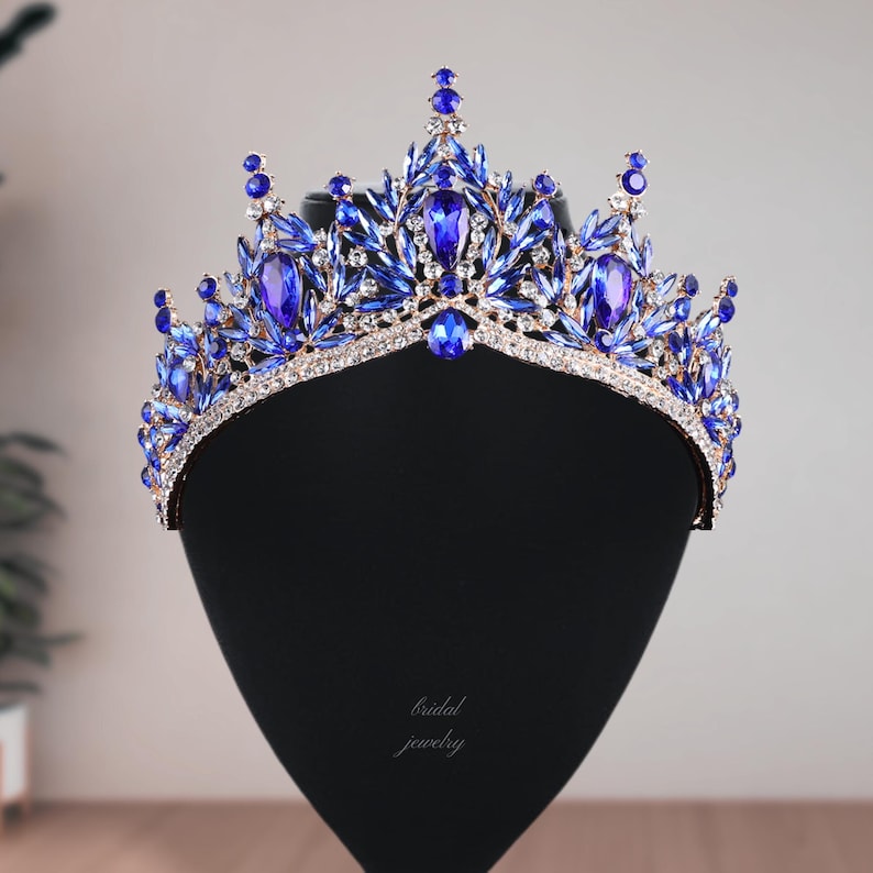May include: A regal blue and silver crown with large, oval-shaped blue gemstones and smaller, round blue accents. The crown is set on a black display stand, with the words "bridal jewelry" in cursive script.